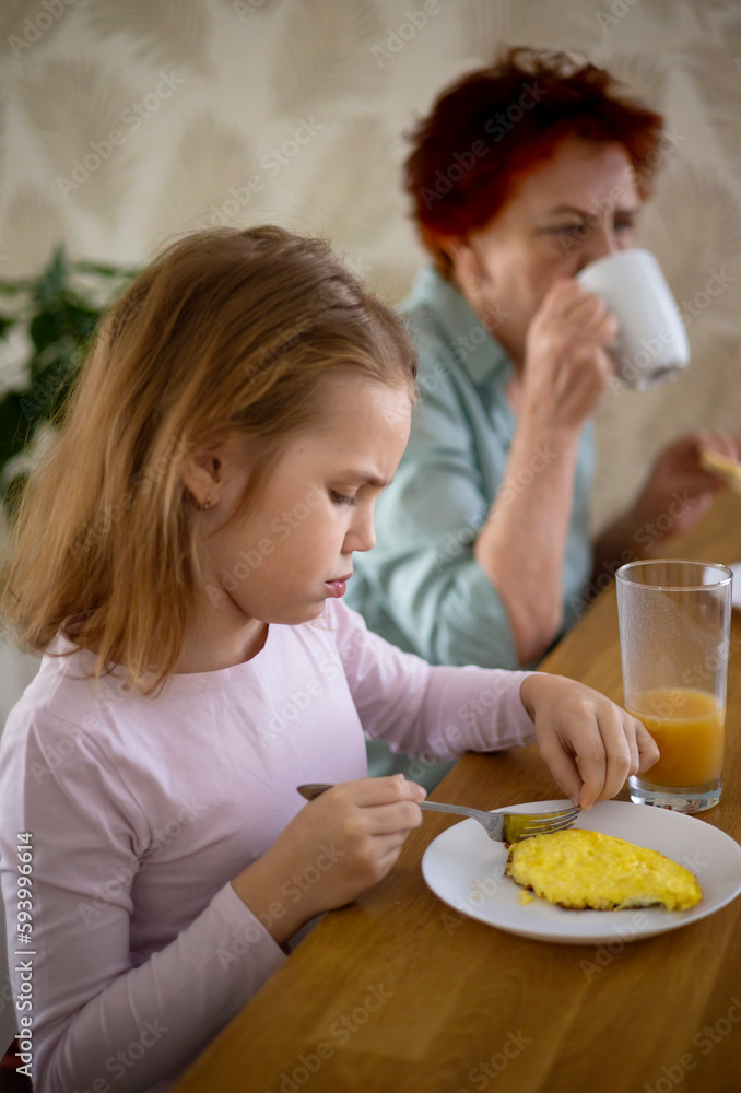 A little blonde girl is having breakfast in the kitchen with her beloved grandmother. The child eats a healthy breakfast with an appetite. Family traditions and friendship between generations.