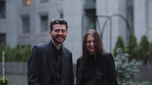 Two young men in strict black suits are laughing while looking at the camera. Business partners pose against the backdrop of a blurred building.