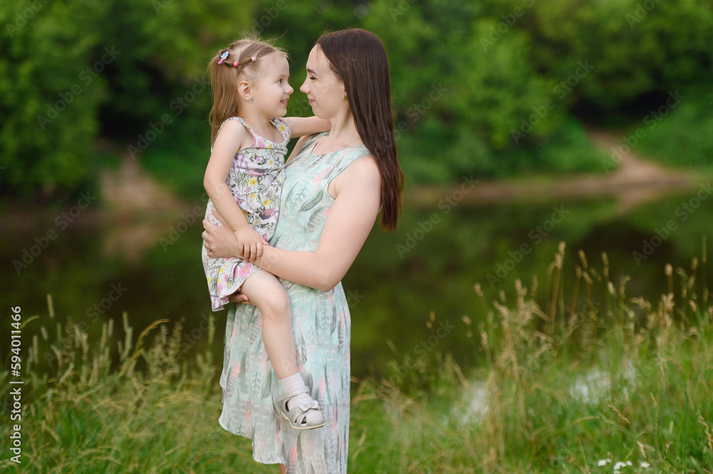 Fototapeta premium Happy mother holding her daughter in her arms, hugging,spending time together,enjoy happy family time. The concept of a happy family, family day, mother's day, countryside vacation, unity with nature.
