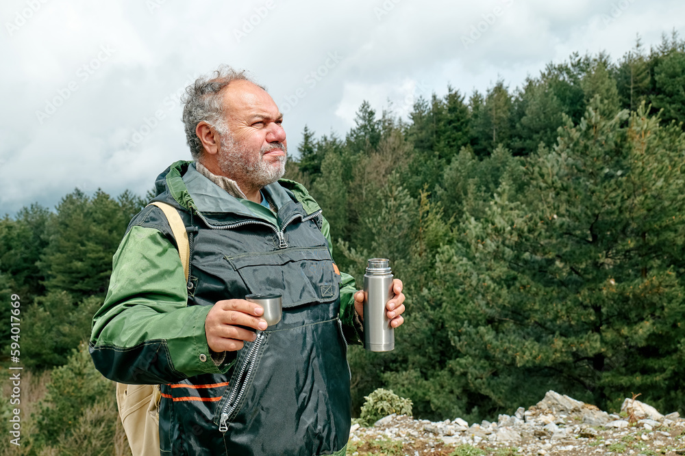 Traveler man hiking in mountain forest, relaxing while drinking a cup of coffee from thermos on panoramic mountain valley.