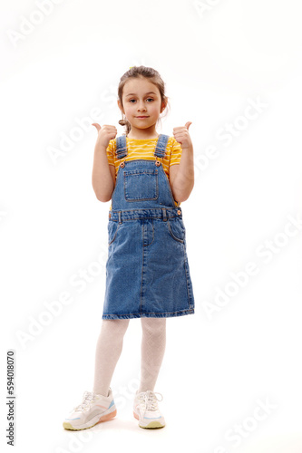 Full-length portrait of Caucasian lovely little girl in casual denim clothes, looking at camera, gesturing with thumbs up, standing over isolated white background. Schoolgirl. Portrait of a smart kid