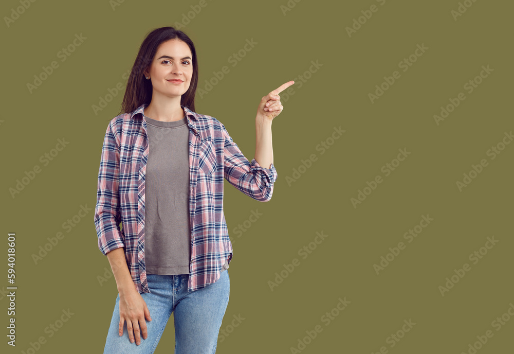 Happy beautiful young woman wearing casual plaid shirt, T shirt and jeans standing isolated on dark green background and pointing to side to show something on blank empty advertising copy space Happy beautiful young woman wearing casual plaid shirt, T shirt and jeans standing isolated on dark green background and pointing to side to show something on blank empty advertising copy space