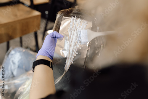 Fotografie Unrecognizable woman hairstylist colorist hold strand of long hair on foil, applying hair color with brush in gloves