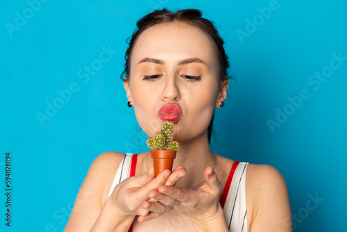 Beautiful young woman kisses a tiny cactus. Isolated on blue background.
