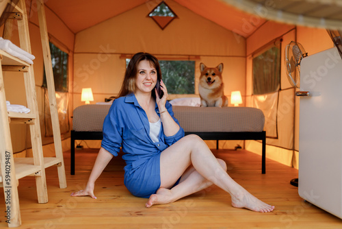 Happy young woman with Welsh Corgi Pembroke dog relaxing in glamping and talking phone. Luxury camping tent for outdoor recreation and recreation. Lifestyle concept
