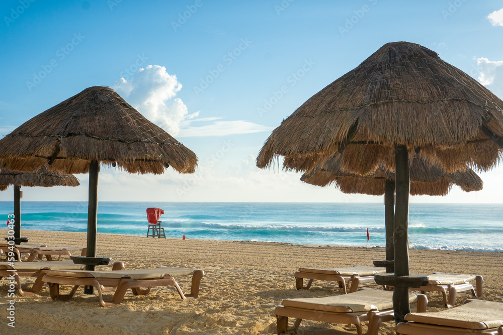 empty lounge chairs with straw umbrellas along the beach with a lifeguard station with red awning in the background. Beach located in the Zona Hotelera (Hotel Zone) in Cancun, Mexico. 
