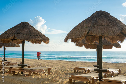 empty lounge chairs with straw umbrellas along the beach with a lifeguard station with red awning in the background. Beach located in the Zona Hotelera  Hotel Zone  in Cancun  Mexico. 