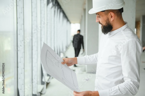 Serious busy young attractive arabic industrial engineer in hard hat with project drawings in factory interior. Professional and industry, builder architect job, worker checking work at plant indoor