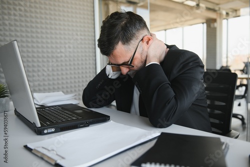 Slika na platnu Worried businessman in dark suit sitting at office desk full with books and papers being overloaded with work