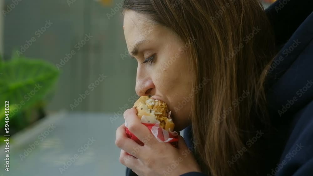Brunette girl 20s in hoodie eating burger in city cafe or city mall. Junk unhealthy food. Concept of meat nutrition. Close up. Slowmotion. Brunette girl 20s in hoodie eating burger in city cafe or city mall. Junk unhealthy food. Concept of meat nutrition. Close up. Slowmotion.