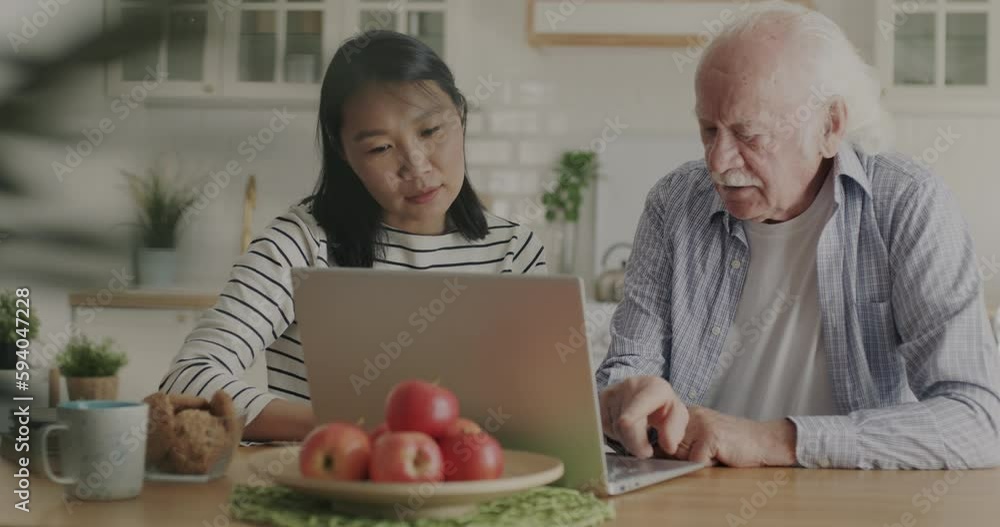Nurse Asian woman teaching elderly man how to use laptop computer helping with gadget indoors in kitchen. Caregiver and old age concept. Nurse Asian woman teaching elderly man how to use laptop computer helping with gadget indoors in kitchen. Caregiver and old age concept.