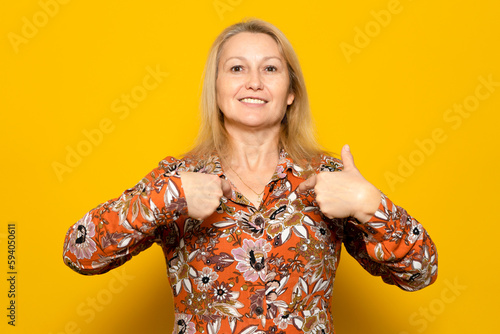 Caucasian blonde woman in her 40s in a patterned dress pointing to herself proud of being the way she is, strong and independent woman isolated over yellow background.