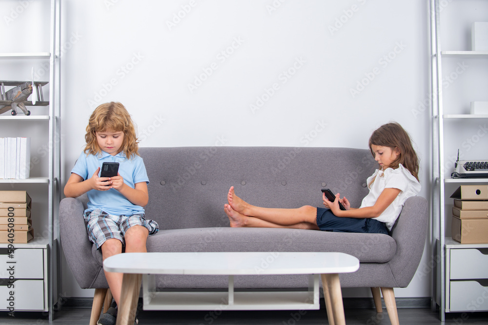 Kid with smartphone at home. Two cute children using online app on mobile phone, making call, watching content, browsing internet, playing video games, sitting on couch in at home alone. Kid with smartphone at home. Two cute children using online app on mobile phone, making call, watching content, browsing internet, playing video games, sitting on couch in at home alone.