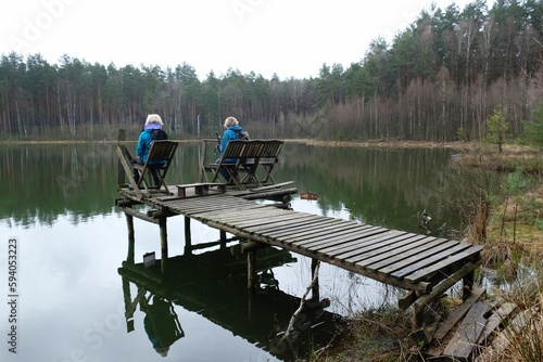 Two women in blue jackets are sitting on a wooden pier by the river on a spring day.