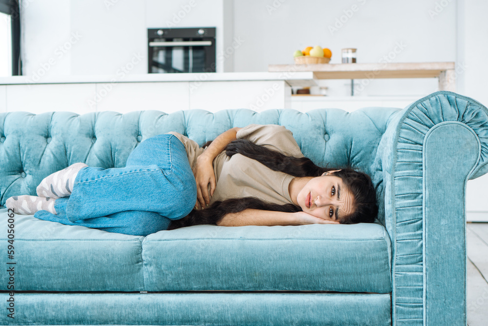 Sick girl with stomach ache menstrual pain and bad feeling lays down on the couch with her legs tucked up Sick girl with stomach ache menstrual pain and bad feeling lays down on the couch with her legs tucked up