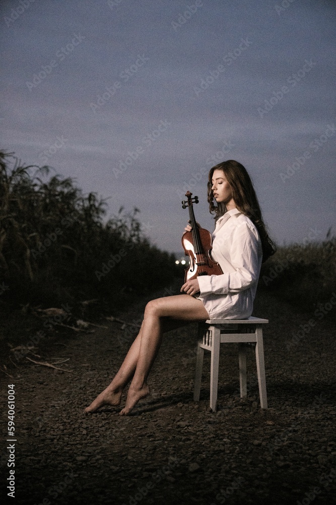 Vertical shot of a young Caucasian girl with a violin sitting on a chair in a field Vertical shot of a young Caucasian girl with a violin sitting on a chair in a field