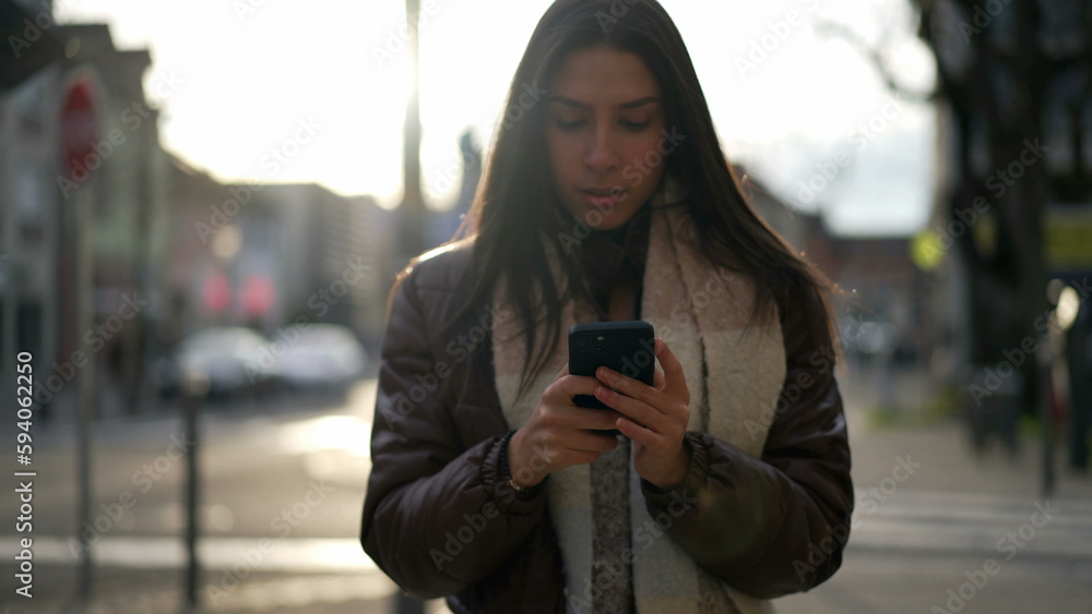 Obraz premium One young adult girl in 20s reading text message on cellphone device while walking outside in city street during afternoon sunlight