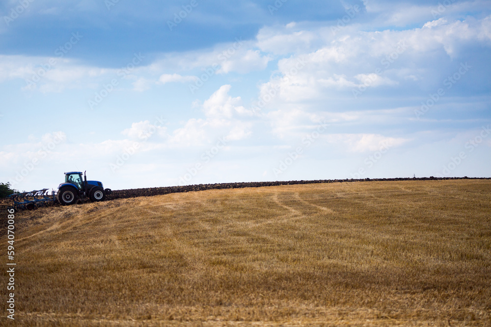 Fototapeta premium A modern blue tractor with an attached reversible plow with a husking roller is plowing a field on which the spring grain crop has just been harvested. Midsummer in central Ukraine.