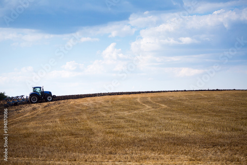 A modern blue tractor with an attached reversible plow with a husking roller is plowing a field on which the spring grain crop has just been harvested. Midsummer in central Ukraine.