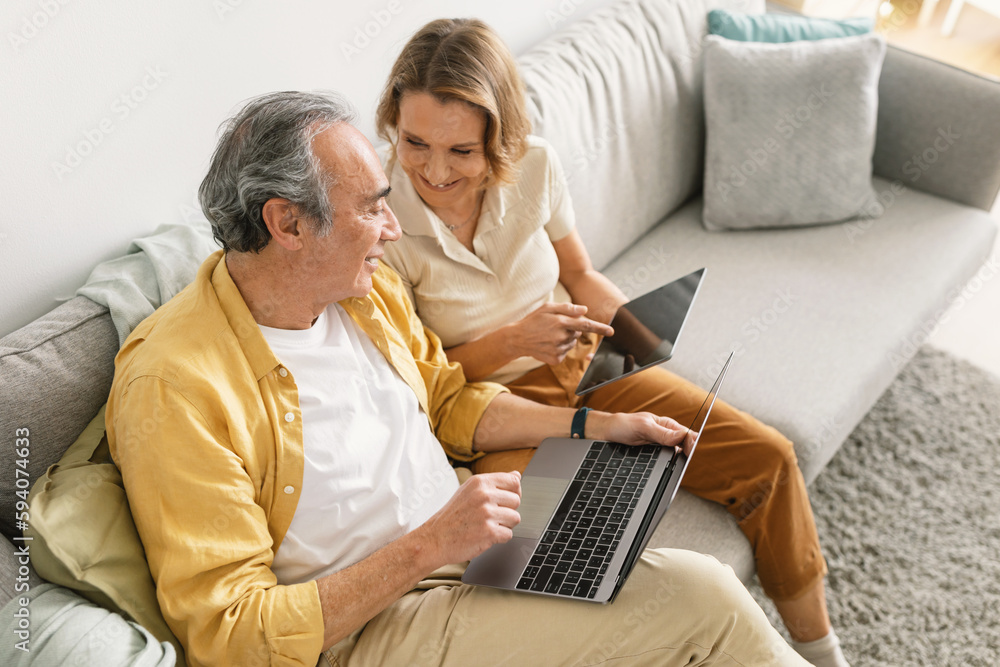 Modern senior couple using laptop computer and digital tablet, sitting on couch at home and talking Modern senior couple using laptop computer and digital tablet, sitting on couch at home and talking