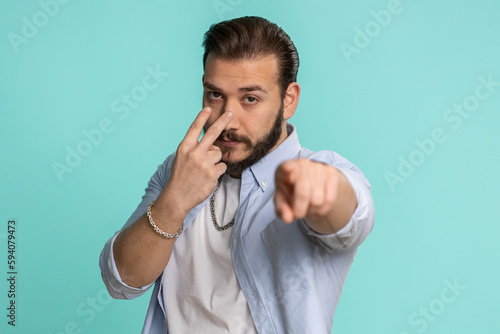 I am watching you. Young confident lebanese man pointing at his eyes and camera, show I am watching you gesture spying on someone. Handsome bearded arabian guy isolated alone on blue studio background