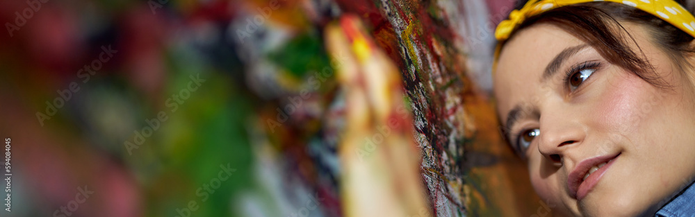 Close up portrait of relaxed young woman, female painter applying paint on canvas with fingers while creating a large modern abstract oil painting