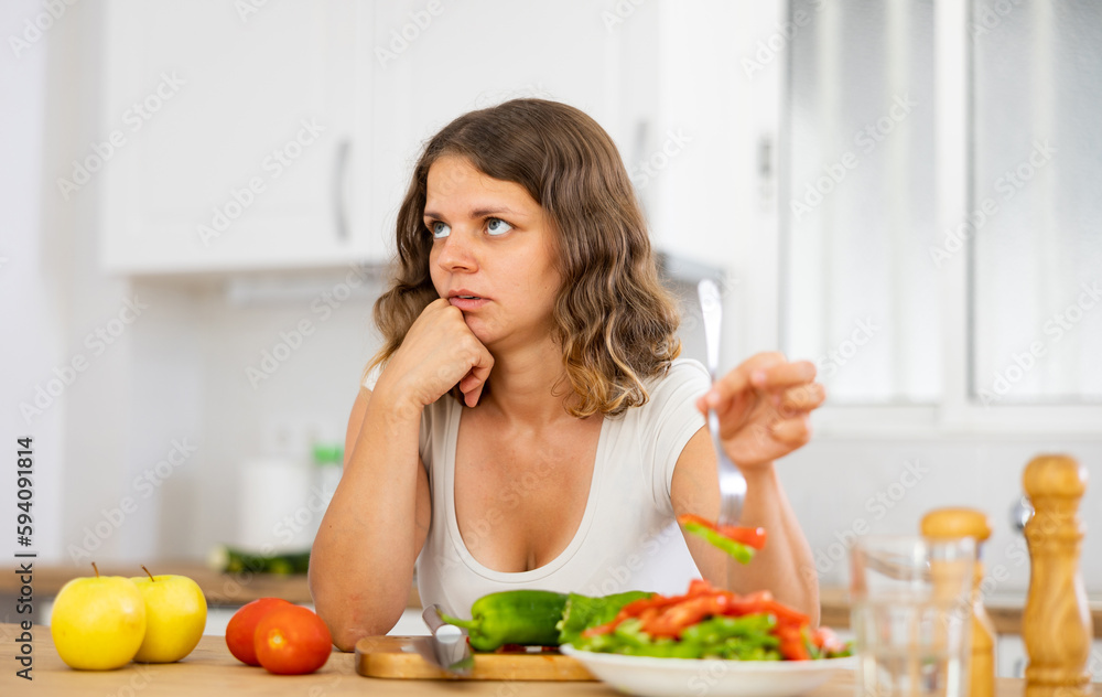 Portrait of displeased woman in nightie eating salad in kitchen at home. Dissatisfied woman having healthy dinner.