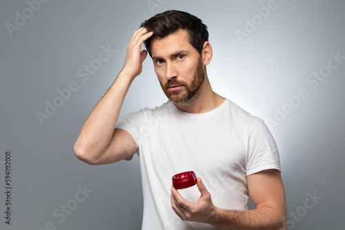 Hair care concept. Confident bearded man holding hair gel and touching his ideal hair, grey studio background photo