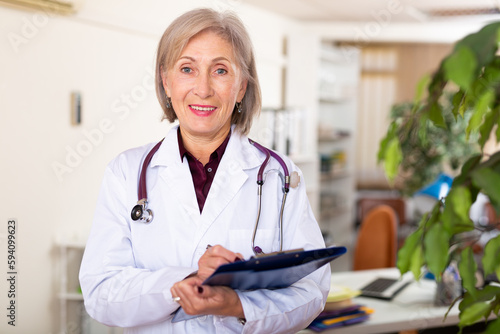 Smiling elderly woman general practitioner in white coat with phonendoscope on neck standing in office with clipboard in hands, writing medical history sheet