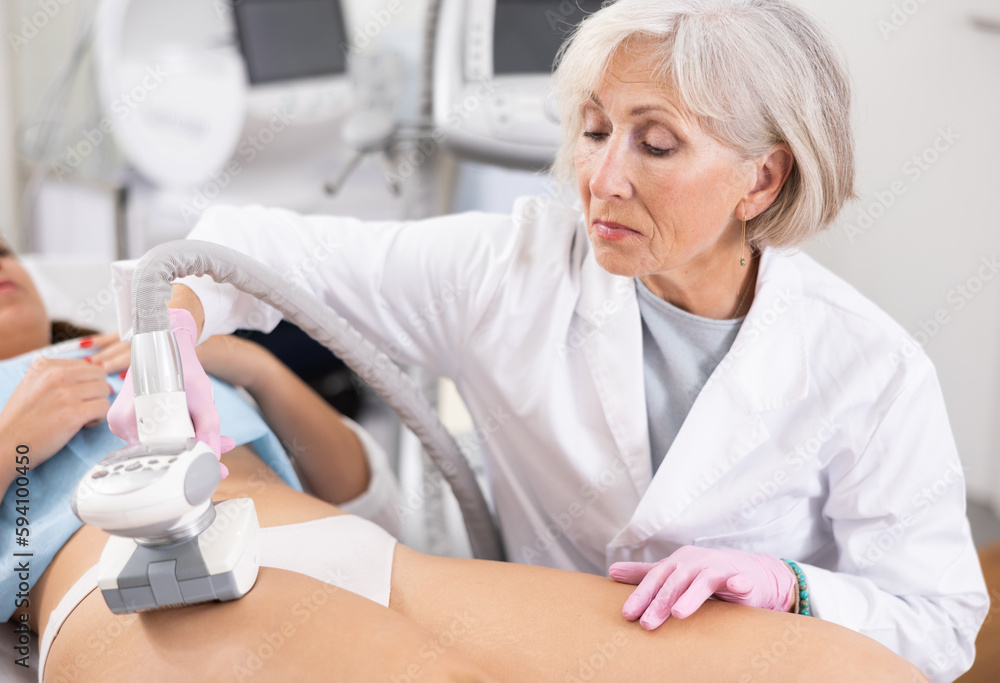 Fototapeta premium Young woman getting ultrasound cavitation body contouring treatment, anti-cellulite therapy, vacuum massage from old female cosmetologist. Hardware cosmetology for body sculpting
