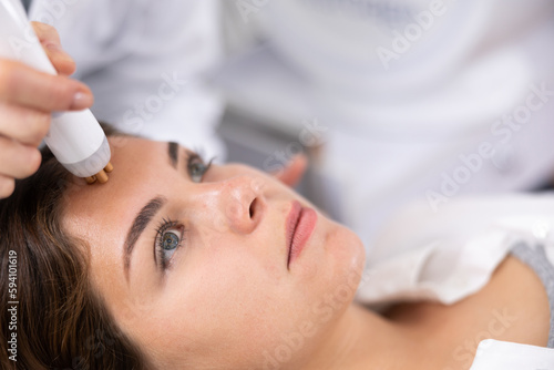 Unrecognizable beautician doctor doing RF lifting procedure for flawless woman face laying in beauty salon. Hardware cosmetology