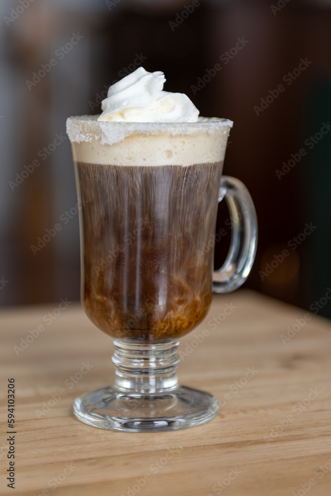 A clear glass mug of Irish coffee with whipped cream and a sugar rim. The tall glass is on a wooden bar at a pub. The white thick cream is falling to the bottom of the hot coffee and strong whisky. A clear glass mug of Irish coffee with whipped cream and a sugar rim. The tall glass is on a wooden bar at a pub. The white thick cream is falling to the bottom of the hot coffee and strong whisky.