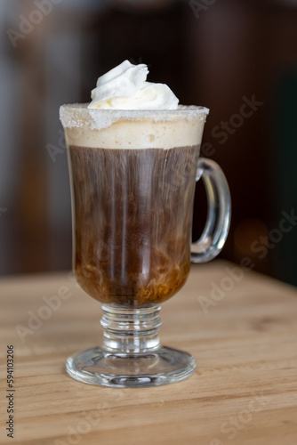 A clear glass mug of Irish coffee with whipped cream and a sugar rim. The tall glass is on a wooden bar at a pub. The white thick cream is falling to the bottom of the hot coffee and strong whisky.