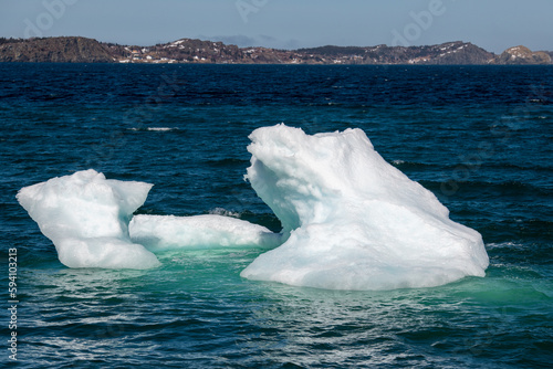 A small iceberg with peaks and craters floating in the cold Atlantic blue ocean. The floe under the water is a teal-green color. Waves are hitting the back of the ice causing a spray over the ice. 