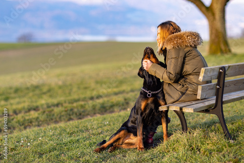 A woman sits on a bench and hugs a Rottweiler on a hill in the morning