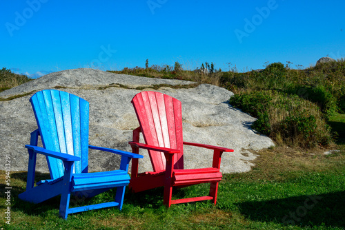 Bright red and blue empty sturdy resin material Adirondack chairs. The furniture is on green lush grass with rocky ground and rolling hills. The terrain is a textured molten pebbly rock surface. 