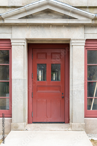A wide vintage red wooden door with two glass pane windows on the top and panels on the bottom. There are two sidelights in the old limestone block building with a decorative limestone overhang.