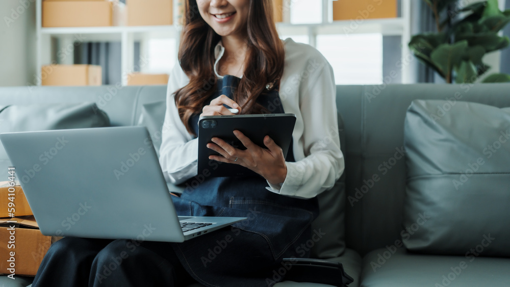 Using tablet pc, Young pretty Asian female influencer small businesses SME owners female entrepreneurs working on receipt box and check online orders, sitting on sofa of home online business. Using tablet pc, Young pretty Asian female influencer small businesses SME owners female entrepreneurs working on receipt box and check online orders, sitting on sofa of home online business.