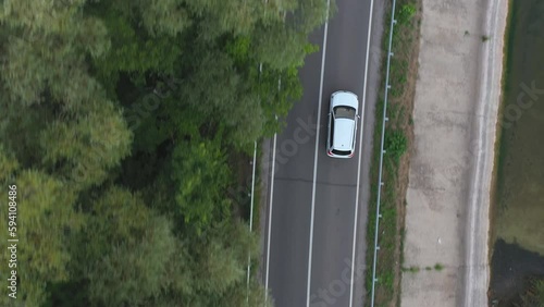 Top view of white car rides through dam route. SUV drives at road near lake on summer day. Flying over the auto moves through bridge of river or pond. Scenic landscape way. Travel concept. Aerial shot
