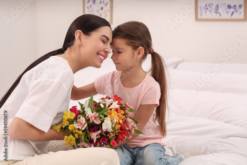 Happy woman with her daughter and bouquet of lilies on bed at home, space for text. Mother's day celebration