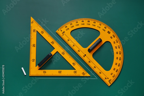 Protractor, triangle and chalk on green chalkboard, flat lay