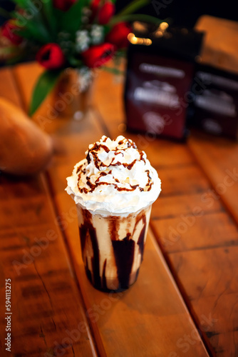 glass of frapuccino with decoration of white cream and chocolate sauce on the wooden table along with packets of caffe and vase photo