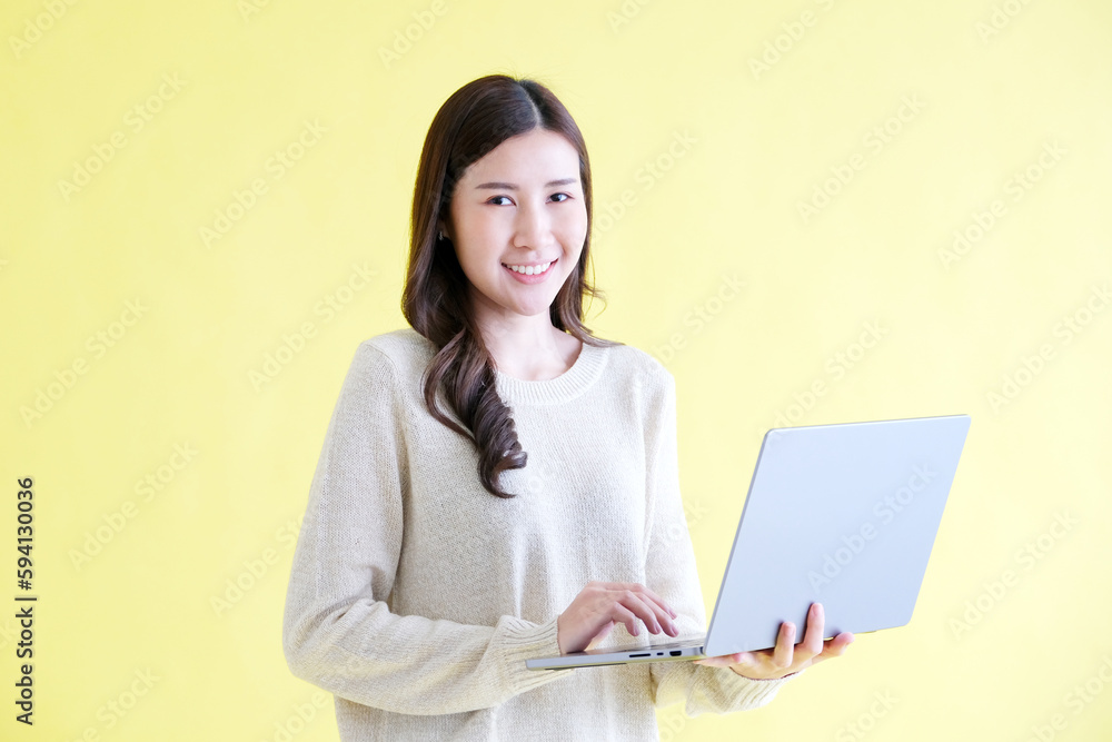 Young asian woman holding laptop computer, smiling and looking at camera while standing over isolated yellow background Young asian woman holding laptop computer, smiling and looking at camera while standing over isolated yellow background
