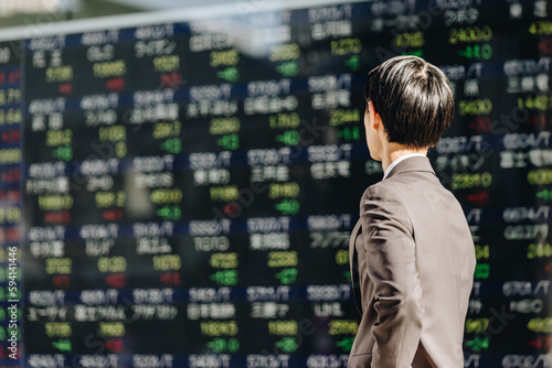 電光掲示板に表示された大量の株の銘柄一覧が表示された株価ボードを眺める30代の日本人の男性の後ろ姿 © chachamal