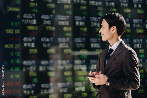 電光掲示板のリアルタイム株価ボードの前でスマートフォンのアプリで株取引をする30代の男性 photo