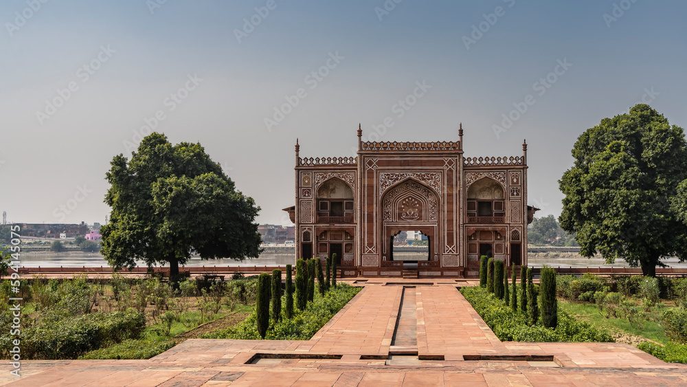 Fototapeta premium The building in the complex of the tomb of Itmad-Ud-Daula, built of red sandstone, by the river. Arches, spires, white ornaments are visible. Green vegetation on the sides of paved footpaths. India. 