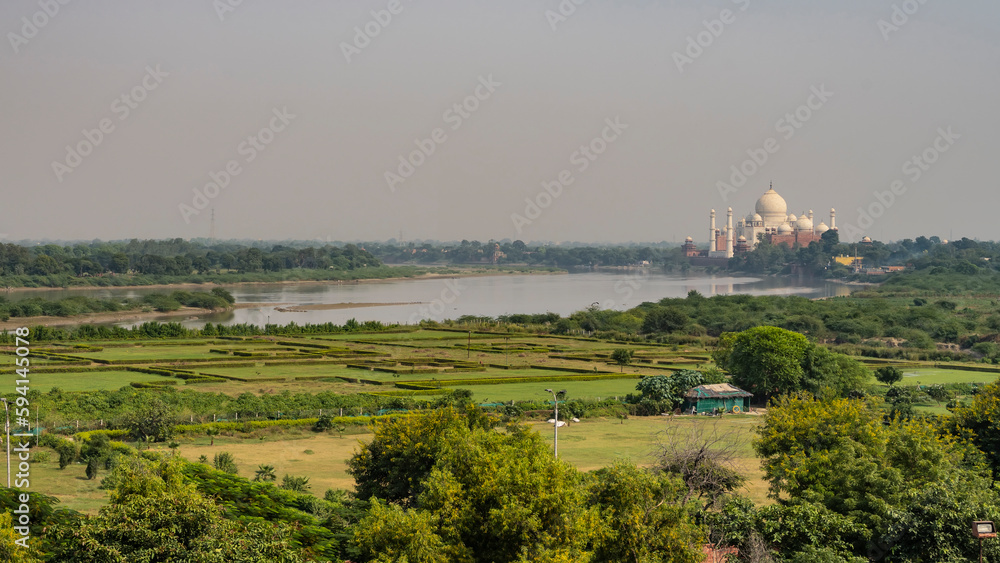 View of the Taj Mahal from the Red Fort. Fields with green vegetation and in the distance, against the sky - a beautiful white marble mausoleum. Reflection in the river. India. Agra
