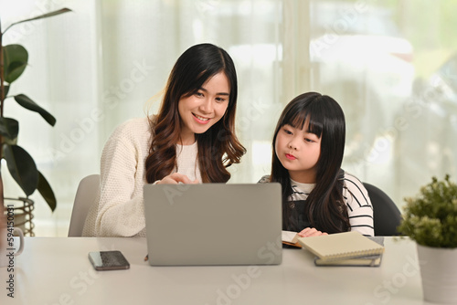 Happy asian mother helping little daughter with primary school home task, sitting at table with books and laptop © Prathankarnpap