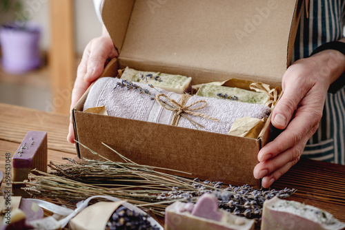 Woman in an apron is holding a craft box with a towel and pieces of lavender natural soap. Handmade gift packaging concept