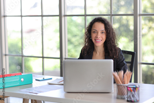 Free Download Attractive professional latin female employee worker sitting, using laptop computer with smart mobile phone at home workplace. Businesswoman working on paperwork while looking at camera and smile - GFXLoad